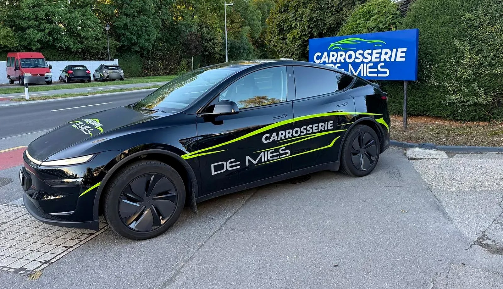 Voiture de prêt aux couleurs de la Carrosserie de Mies devant le panneau de l'atelier