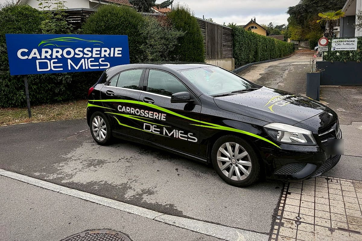 Mercedes Classe A noire aux couleurs de la Carrosserie de Mies, devant le panneau de l’atelier
