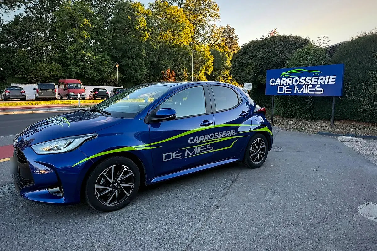Toyota Yaris bleue aux couleurs de la Carrosserie de Mies, devant l’enseigne